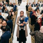 قصة زفاف مهدوم وعدالة بتتحققوتمسك بيدها الأخرى ملفاً أزرق كبيراً.  جميع الحاضرين بملامح مصرية الجالسين على الطاولات يلتفتون إليها بنظرات صدمة وذهول وهمس. في الخلفية البعيدة تظهر منصة الزفاف (الكوشة) وعليها العروس والعريس بملامح مصرية . الإضاءة عالية والتفاصيل حادة وواقعية جداً، نمط دراما اجتماعية."